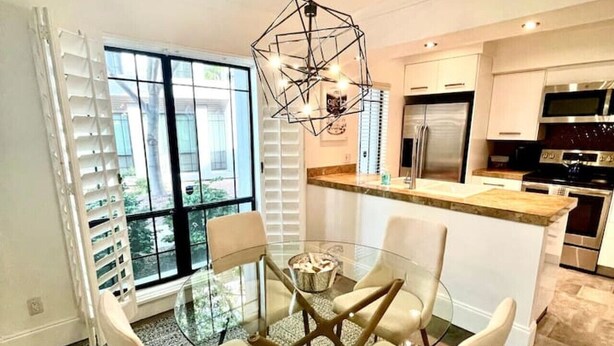 Uma cozinha moderna com uma mesa de jantar de vidro, cadeiras brancas e uma luminária pendente geométrica.