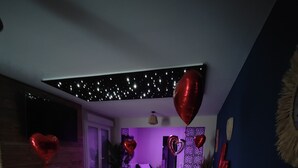 Eine runde Sitzecke mit einem weißen Sofa, einem kleinen Holzschemel und einem Topfpflanzen. Auf dem Boden liegen herzförmige Ballons und eine Anordnung aus Rosenblüten in Herzform.