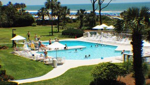 A pool area with lounge chairs and umbrellas, surrounded by greenery and a view of the ocean. - Ocean One 121 - Oceanfront 1st Floor Condo (Hilton Head Island)