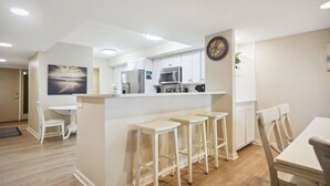 A modern kitchen with white cabinets, a microwave, and a refrigerator. There are three bar stools and a dining table with chairs. - Ocean One 121 - Oceanfront 1st Floor Condo (Hilton Head Island)