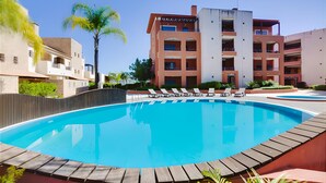 Una piscina con sillas de descanso, rodeada de jardines con palmeras y un edificio de varios pisos al fondo.
