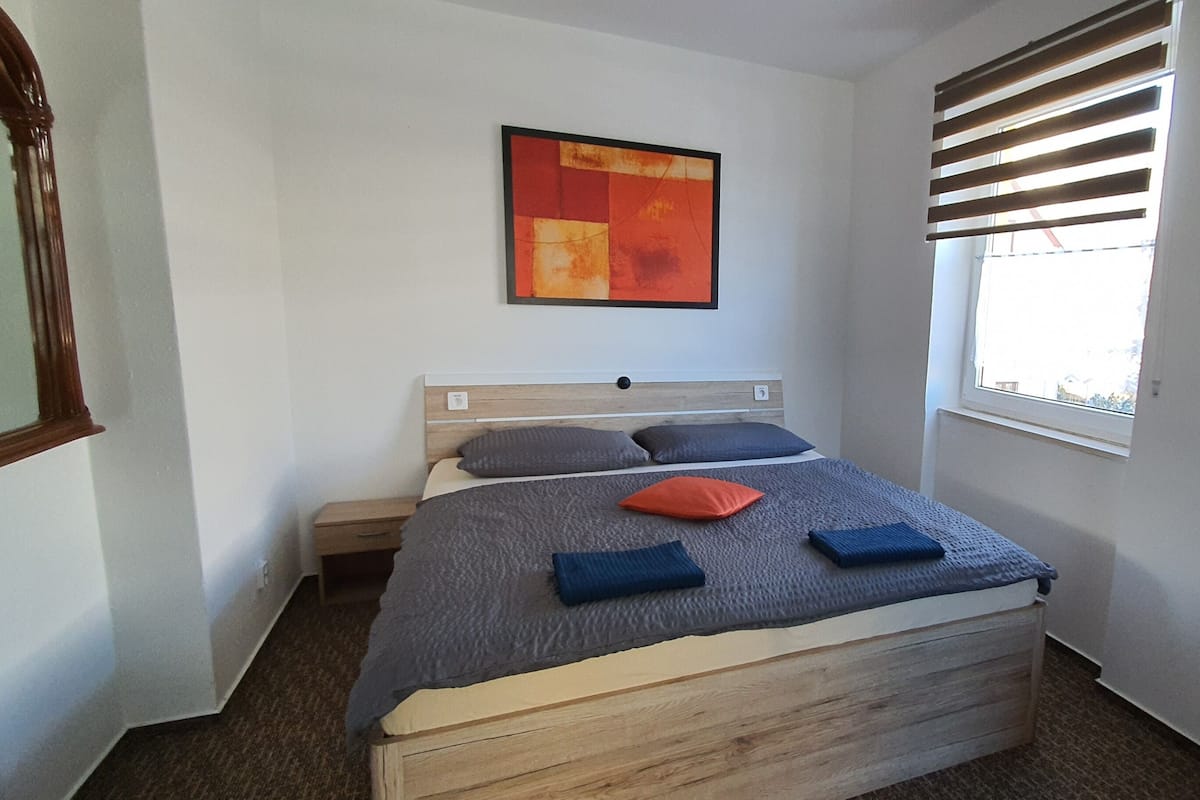 A bedroom with a bed, a window with blinds, a wall-mounted artwork, and a mirror.