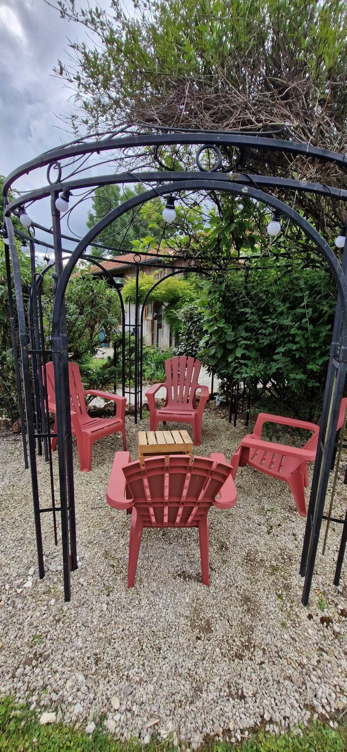 Un gazebo con sedie di plastica rossa e un tavolo di legno, circondato da ghiaia e vegetazione.