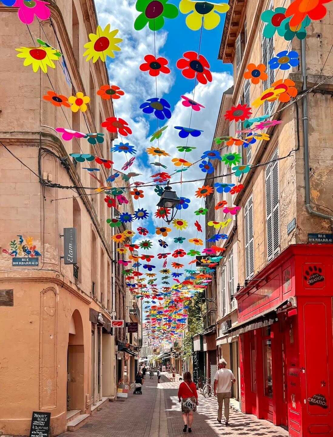 Een smalle straat met kleurrijke papieren bloemen die erboven hangen, gebouwen aan weerszijden en mensen die lopen.