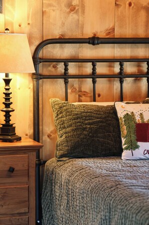 Un letto di legno con testiera in metallo, un cuscino verde con texture, un cuscino bianco con motivo alberi e una lampada su un comodino di legno.