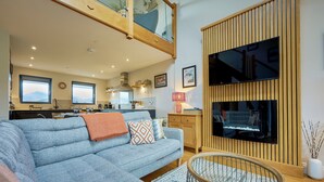 Un salon moderne avec un canapé gris, des éléments décoratifs en bois, un téléviseur fixé sur un panneau en bois, et une cuisine en arrière-plan.