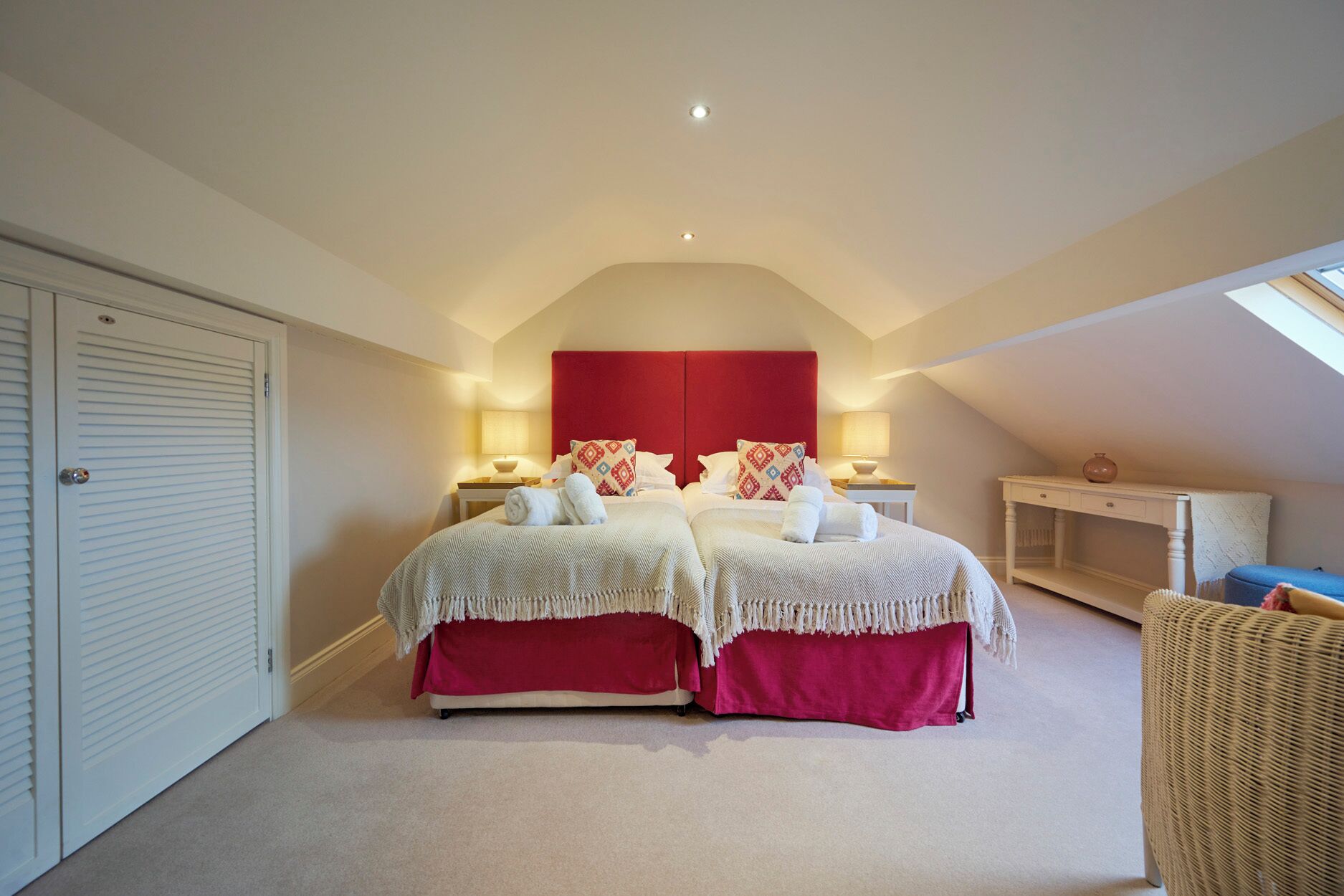 A bedroom with two beds, a red headboard, bedside lamps, a white wardrobe, and a small table with a vase.