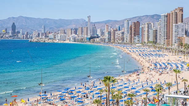 Un paysage urbain côtier avec une plage bordée de parasols bleus et de palmiers.