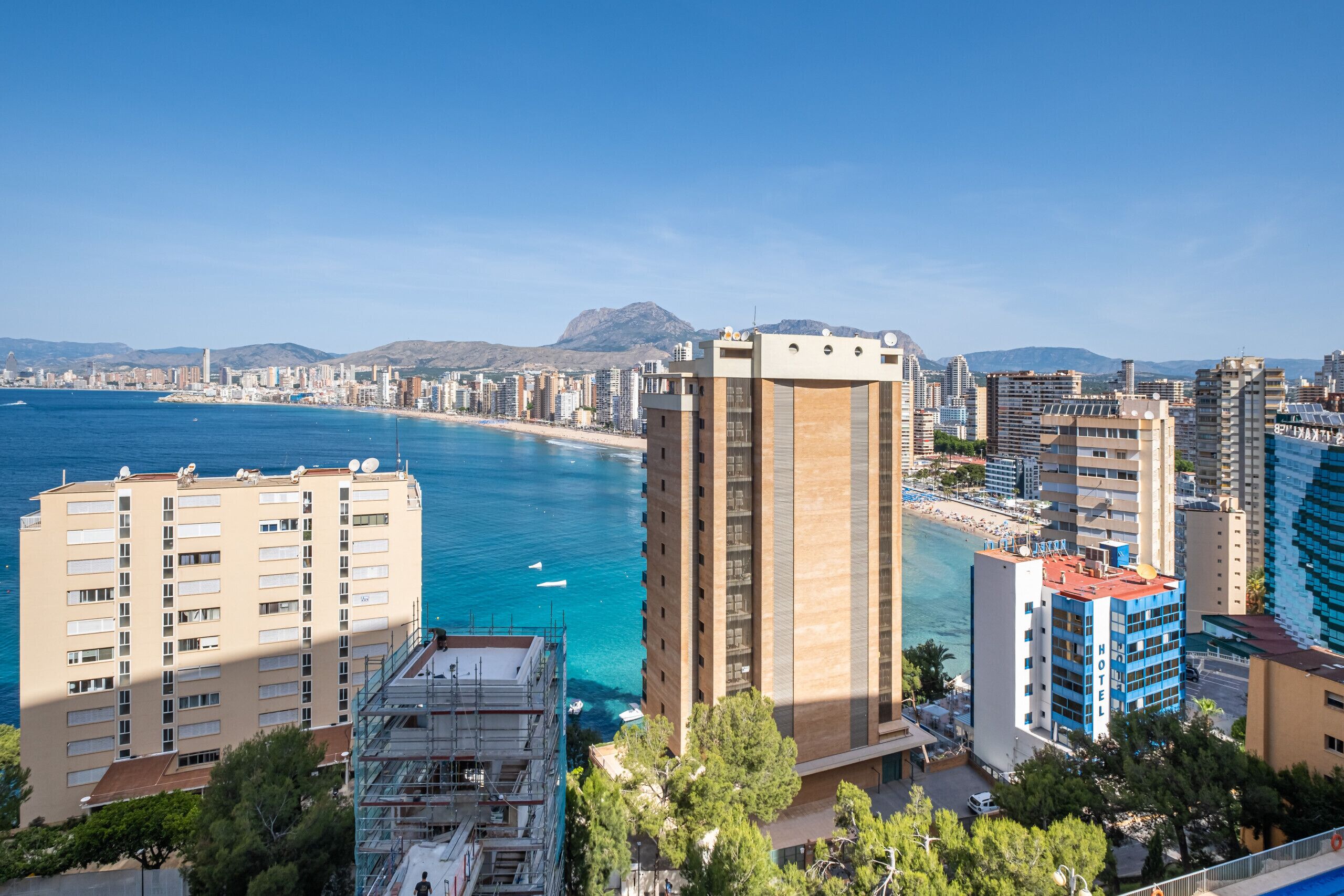 A coastal cityscape with high-rise buildings, a clear blue sea, and a mountainous backdrop.