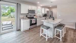A modern kitchen with white cabinetry, a central island with bar stools, stainless steel appliances, and a sliding glass door leading to a patio area. - Spacious w/ all King Beds, 15 min from Beach + Downtown, Pup Friendly! (Wilmington)