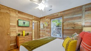 Une chambre avec des murs en bois, un lit avec un matelas blanc, un ventilateur de plafond, un téléviseur fixé au mur et une fenêtre donnant sur un paysage verdoyant.