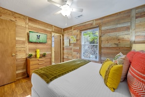 A bedroom with wooden walls, a bed with a white mattress, a ceiling fan, a TV on the wall, and a window with a view of greenery.