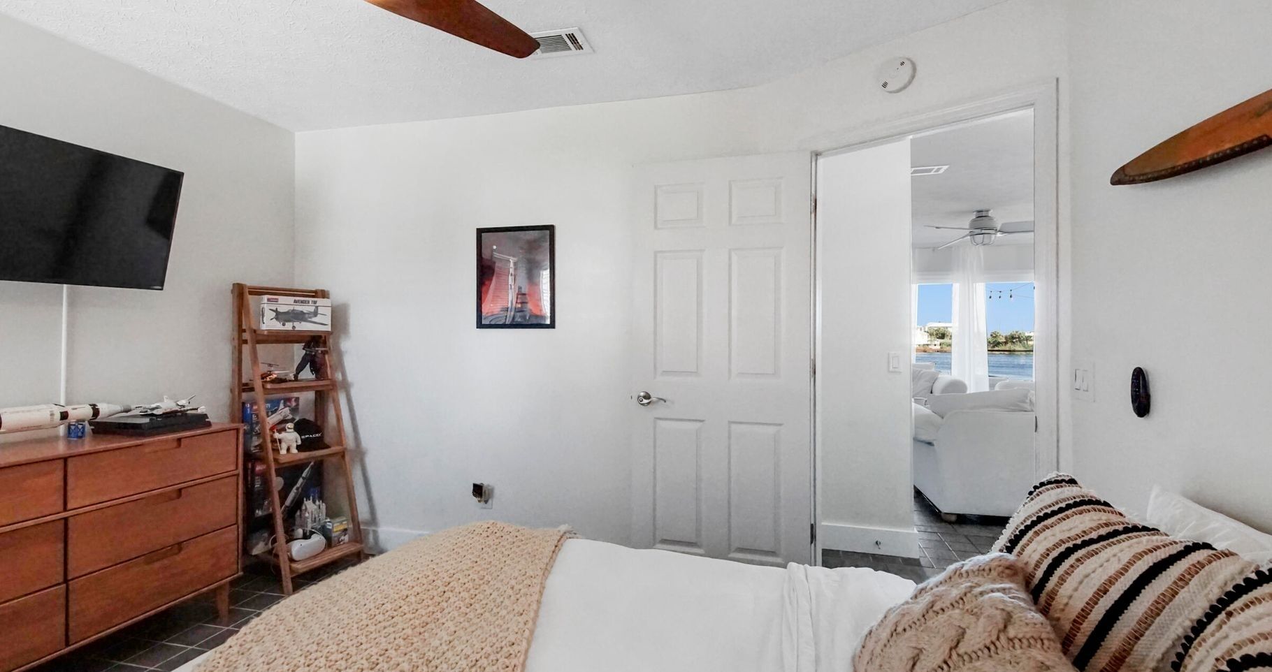 A bedroom with a bed, a TV, a wooden dresser, a shelf with toys, and a door leading to a balcony with a view of the sea.