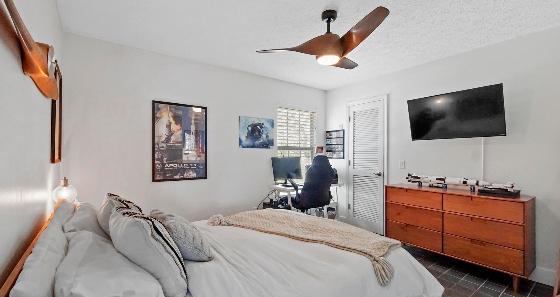 Une chambre à coucher avec un lit, un bureau avec un ordinateur, une télévision et un ventilateur de plafond.