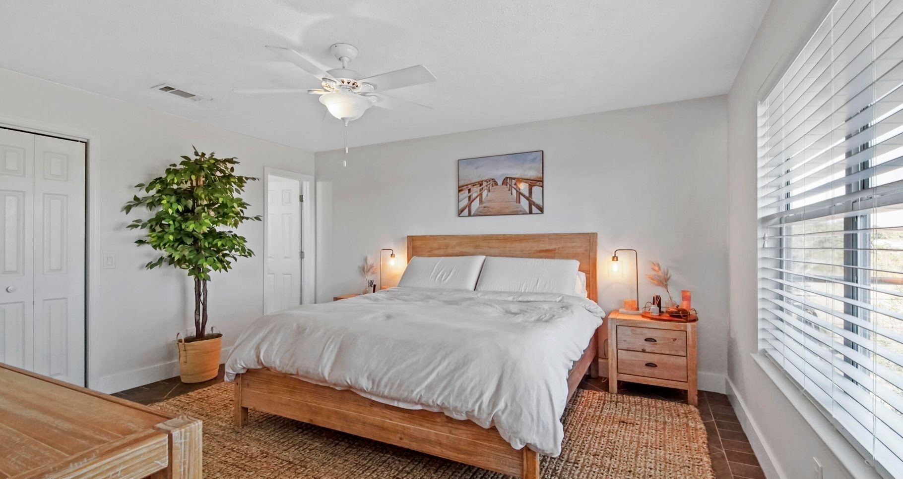 Une chambre avec un lit en bois, un ventilateur de plafond, une plante en pot, une table de chevet avec une lampe et une fenêtre avec des stores.