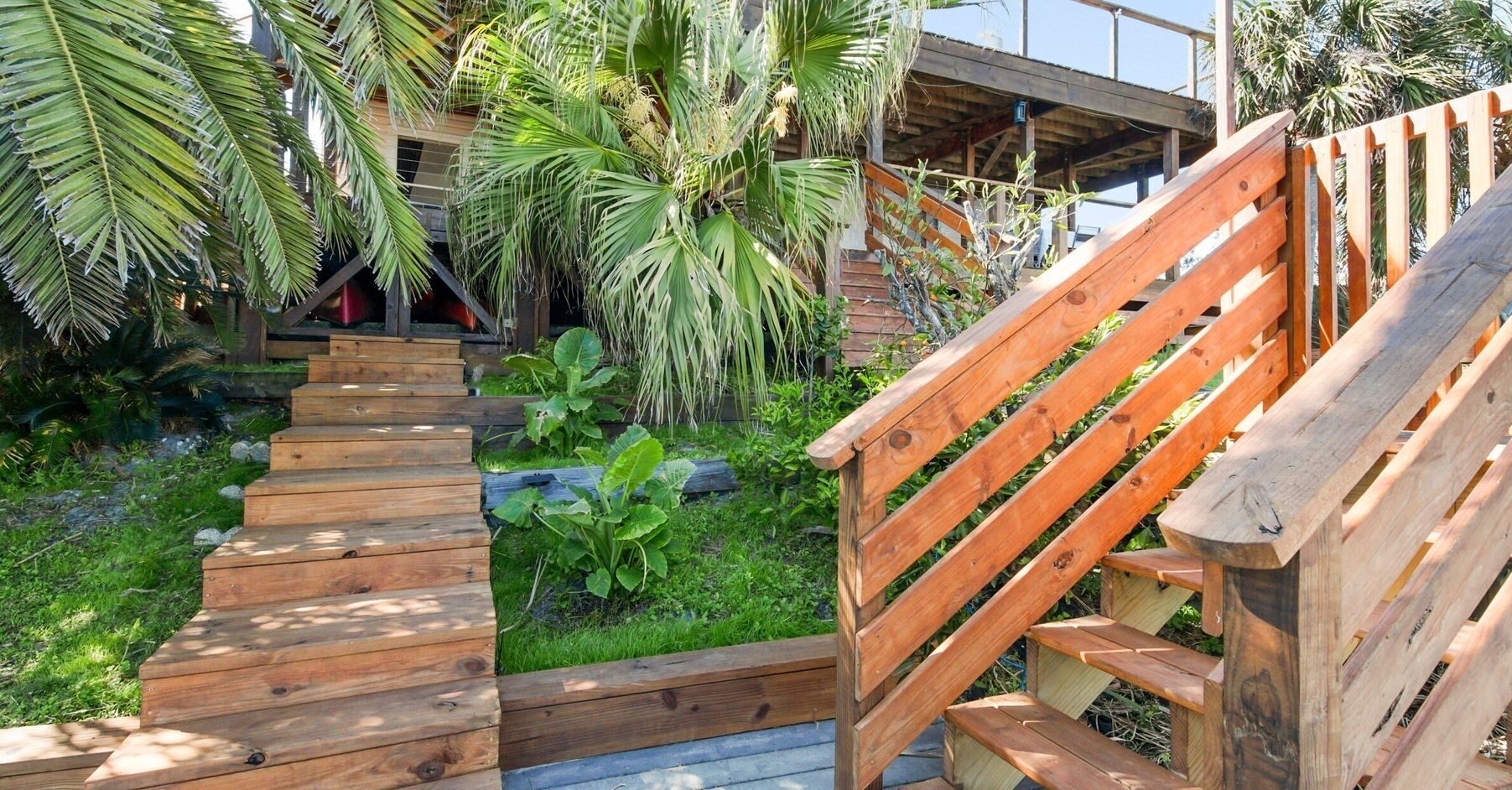Una escalera de madera que conduce a una terraza de madera rodeada de abundante follaje y palmeras.