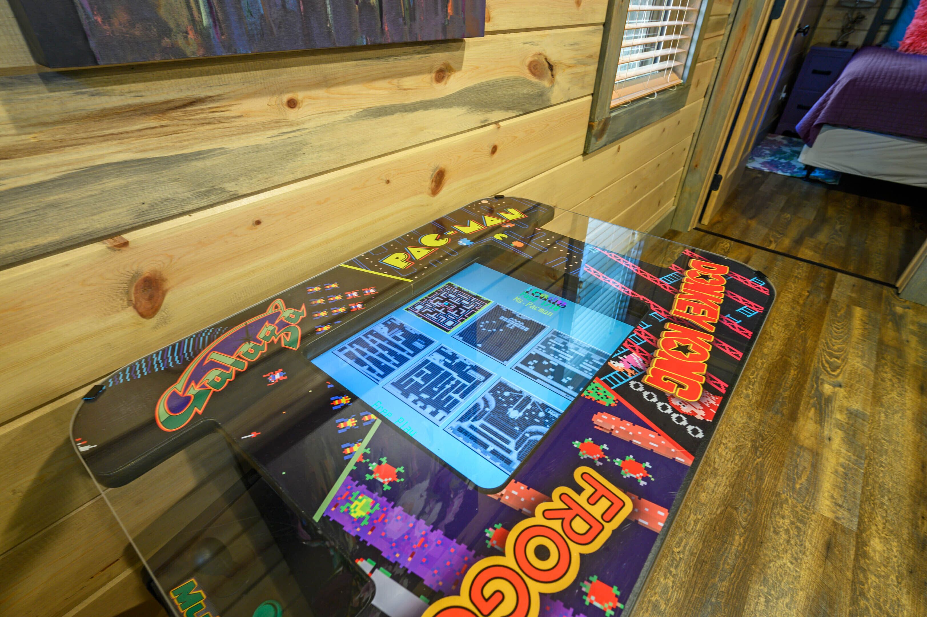 Ein Tisch mit Glasplatte, dessen Oberfläche ein farbenfrohes Spielbrettdesign zeigt, Holzwände und ein Bett im Hintergrund.