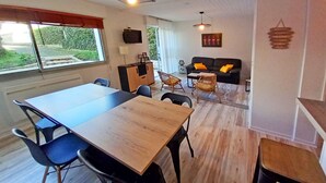 A dining area with a wooden table and chairs, a sofa with cushions, a TV mounted on the wall, and a view of greenery outside. - APARTMENT FACING FONCILLON BEACH (Royan)