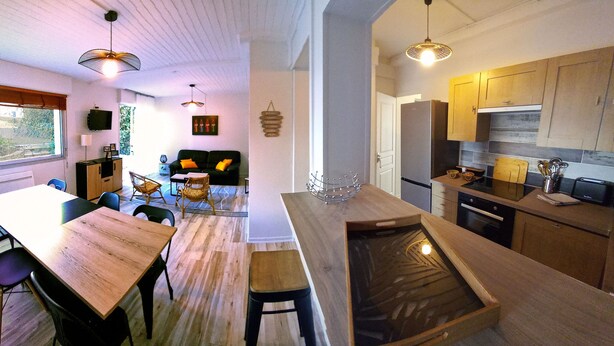 A modern kitchen with wooden cabinets, a dining area with a wooden table and chairs, and a view of the outdoors through large windows. - APARTMENT FACING FONCILLON BEACH (Royan)