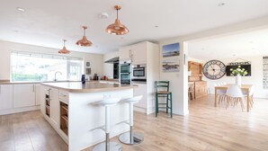 A modern kitchen with white cabinets, a central island with bar stools, copper pendant lights, and a dining area with a wooden table and chairs. - Flaad House (Wadebridge)