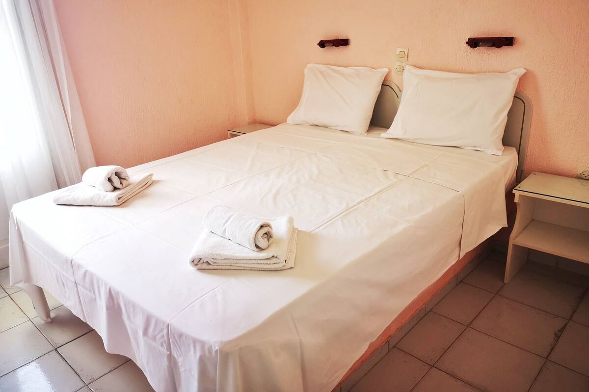 A neatly made bed with white linens and pillows, a folded towel on the bed, and a small bedside table with a lamp.
