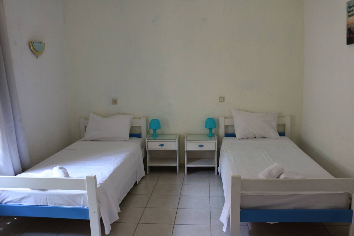 A room with two single beds, each with a white mattress and blue headboard. There are two bedside tables with lamps, and the room has white walls and a tiled floor.
