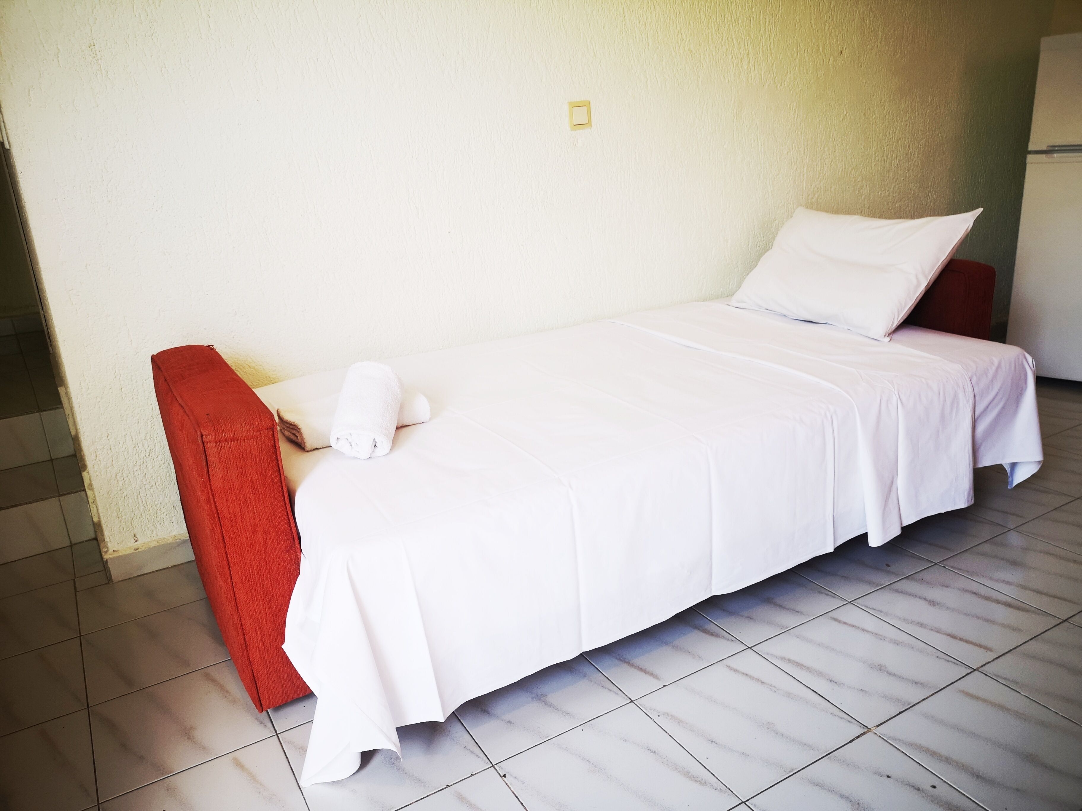 A single-bed room with a white bedspread, a red bench, and a white pillow.