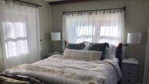 A bedroom with a large bed, two bedside tables with lamps, sheer curtains, and a wooden headboard.