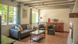 Ein Wohnzimmer mit einer Couch, Sesseln, einem Couchtisch und einem Heizkörper. Es gibt Fenster mit Vorhängen, ein Gemälde an der Wand und eine Holzkabine.