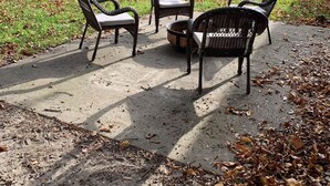 Um pátio com três cadeiras de vime e uma mesa pequena, cercado por árvores e gramado.