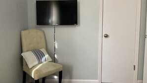 Um quarto com uma poltrona bege, uma televisão de tela plana preta fixada na parede e uma porta branca.