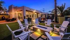 Un patio moderno al aire libre con sillas Adirondack blancas, una chimenea y luces colgantes.