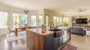 Una cocina moderna con una isla central, un comedor con sillas, un televisor grande y un ventilador de techo.