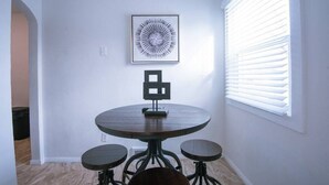 Un coin repas avec une table ronde et quatre chaises, une œuvre d’art abstraite encadrée au mur et une fenêtre avec des stores.