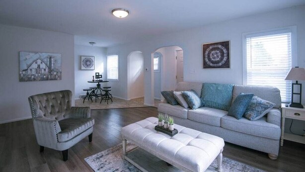 Un salon spacieux avec un canapé gris, un fauteuil à capitons et un pouf blanc. On aperçoit à l’arrière-plan un coin repas avec une table et des chaises.