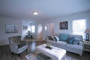 Un salón amplio con un sofá gris, un sillón capitoné y un reposapiés blanco. En el fondo, hay un comedor con una mesa y sillas.