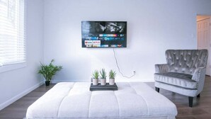 Un salon avec un téléviseur à écran plat fixé au mur, un fauteuil à capitons gris, un pouf blanc et trois plantes en pot.
