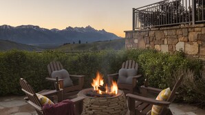 A stone fire pit with a fire, surrounded by wooden chairs with cushions, set on a patio with stone flooring, overlooking a mountainous landscape. - Outpost: Teton Retreat - Expansive Teton Views (Jackson)