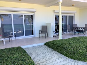 A patio area with a table and chairs, a small table with a vase, and a view of the ocean through large sliding doors. - Castaways Cove Unit 122  Beach Front direct access (East End)