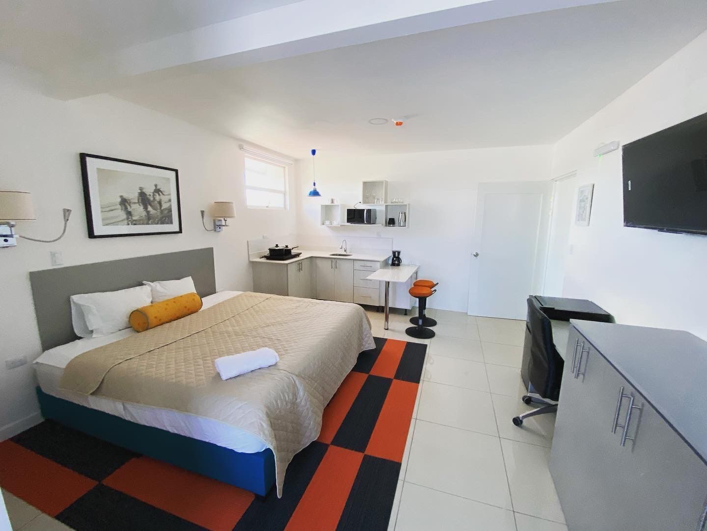 A compact hotel room with a bed, a small kitchenette, a desk, and a TV.
