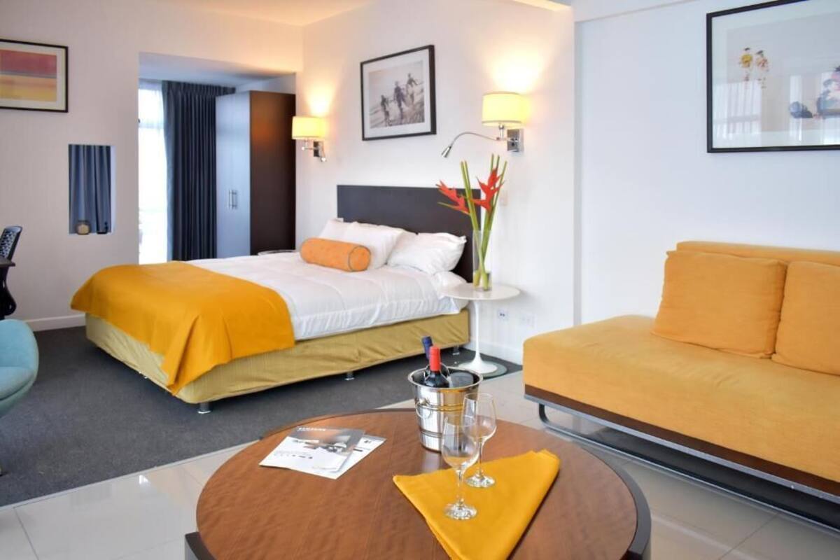 Een hotelkamer met een bed, een bank, een kleine tafel met een glas en een fles, en een vaas met bloemen.