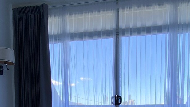 A hotel room with a large bed, a window with sheer curtains, and a view of the cityscape. - Apartahotel Altos de la Sabana (San José)
