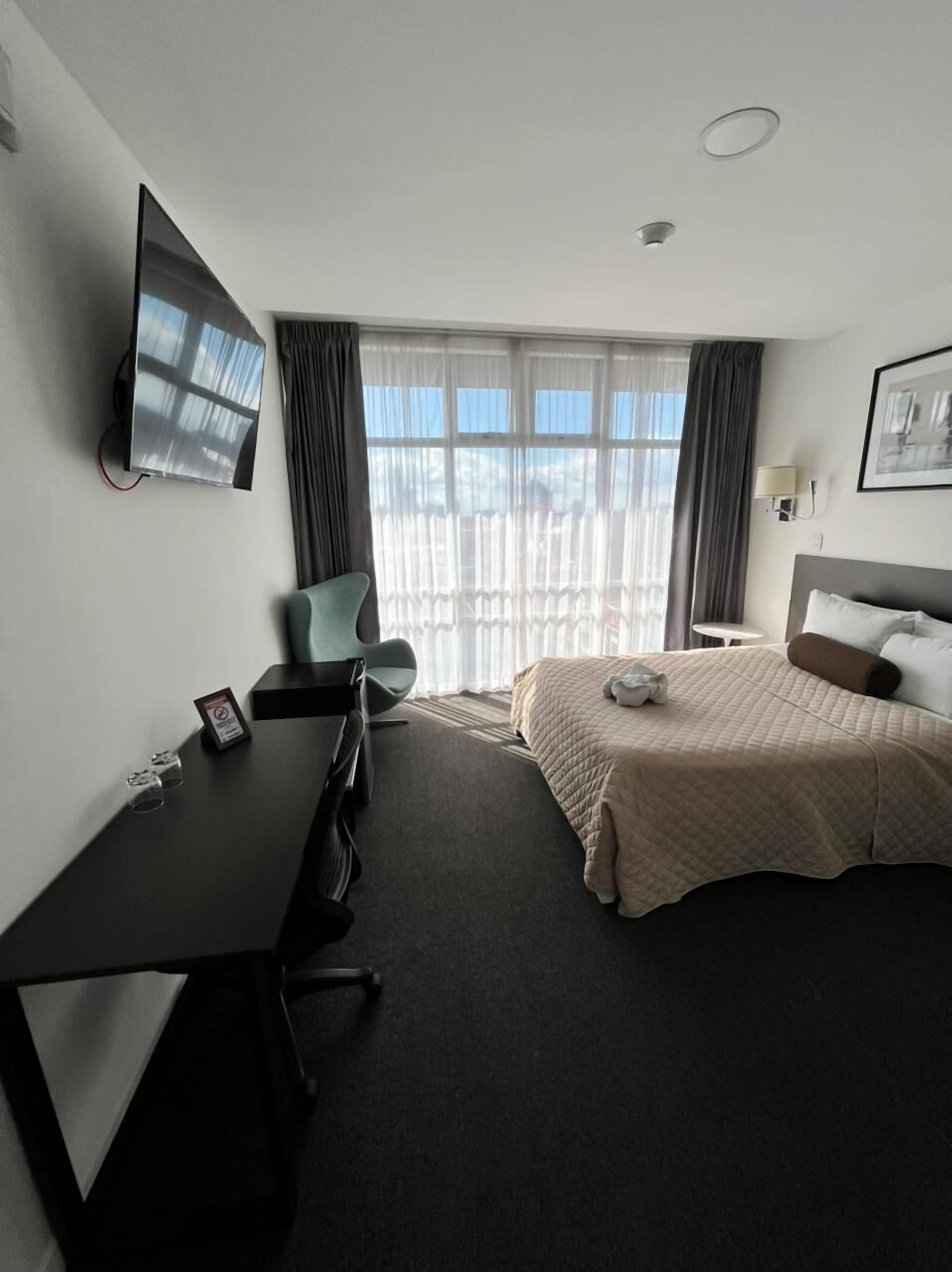 A hotel room with a bed, a desk, a chair, a TV mounted on the wall, and a window with curtains.