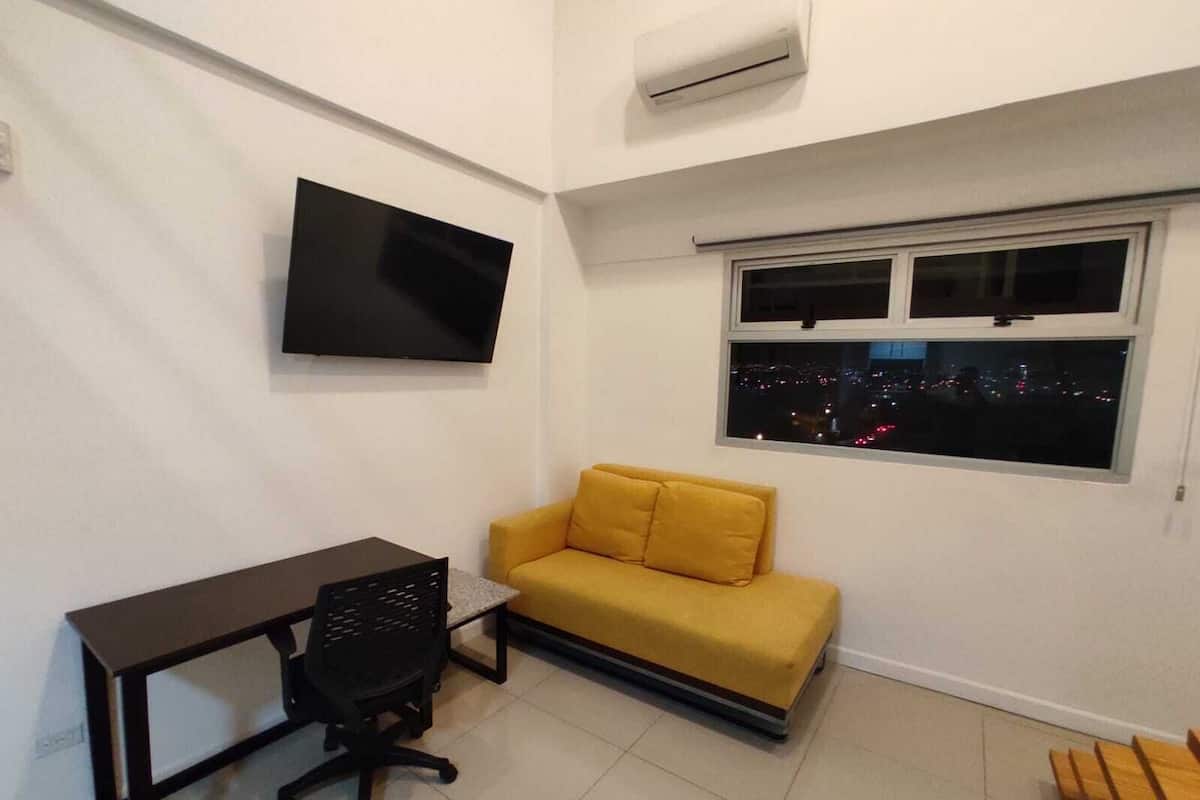 Een kleine, moderne kamer met een bureau, een gele bank, een televisie en een raam met uitzicht op de stad.