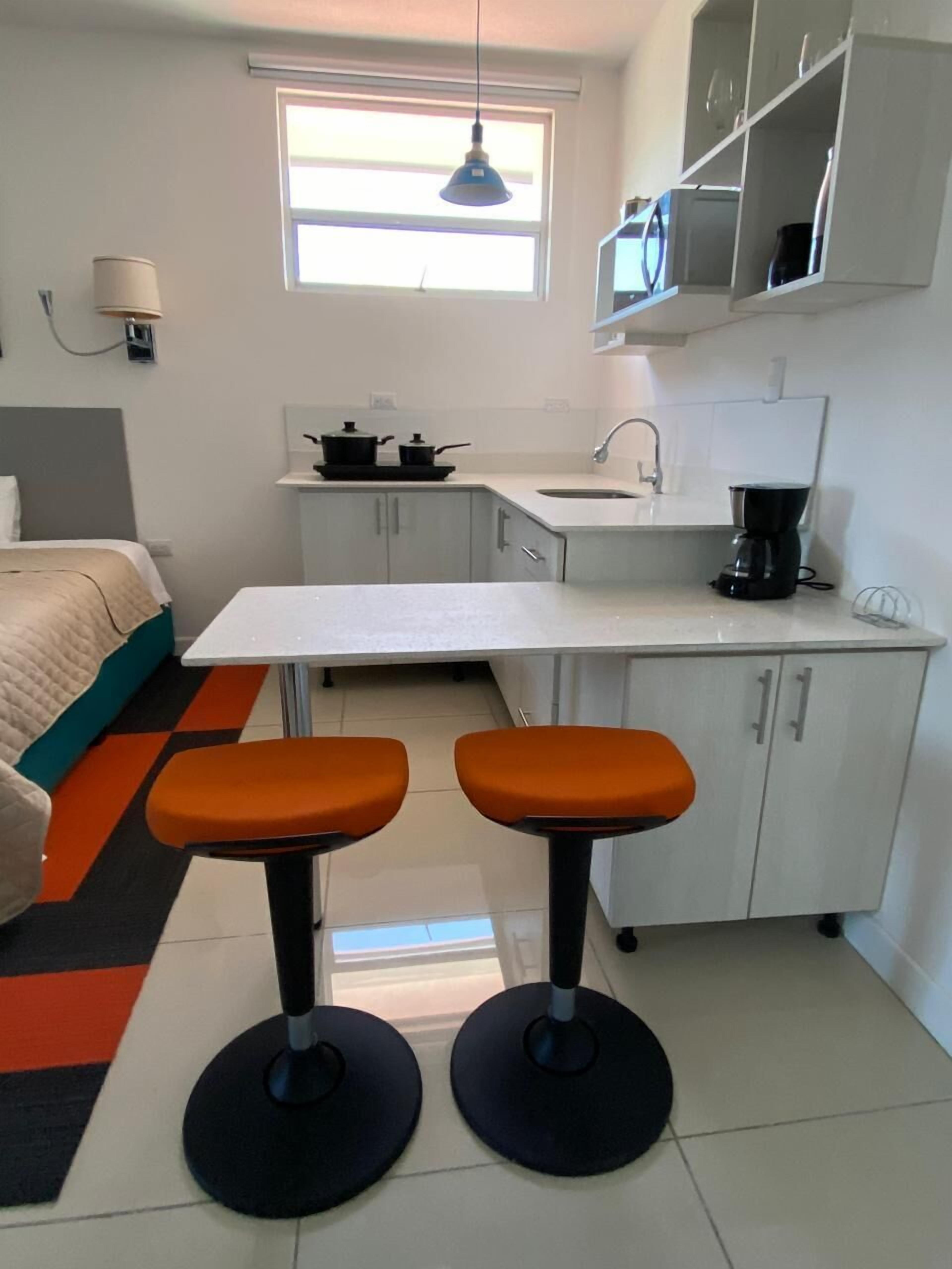 A compact kitchen with a white countertop, two stools, a sink, and a window above the sink.