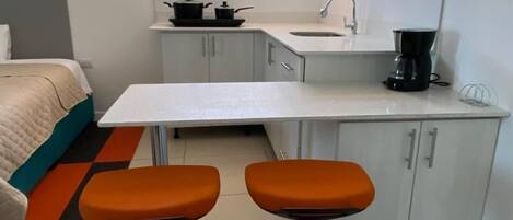 A compact kitchen with a white countertop, two stools, a sink, and a window above the sink.