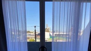 A bedroom with a bed, a window with sheer curtains, and a view of buildings outside. - Apartahotel Altos de la Sabana (San José)