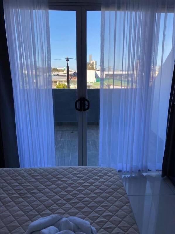 Une chambre à coucher avec un lit, une fenêtre ornée de rideaux légers et une vue sur les bâtiments environnants.