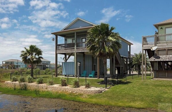 A two-story house with a balcony, surrounded by palm trees and a sandy area.