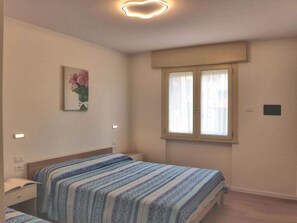 Ein Schlafzimmer mit zwei Betten, einem Fenster mit Vorhängen, einer Wandleuchte und einem Blumenbild in einem Rahmen.